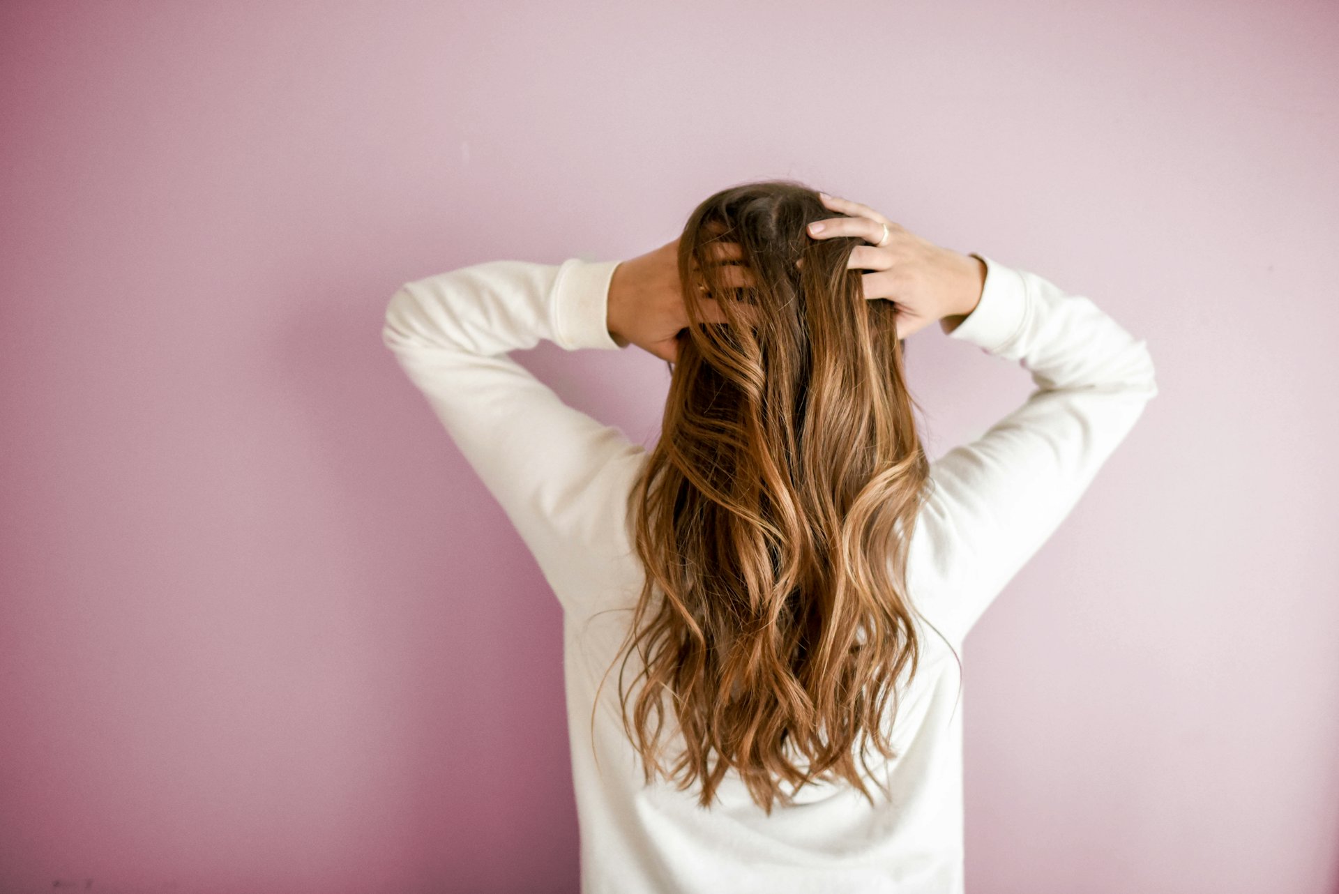 Femme de dos, cheveux longs ondulés aux reflets miel, sur un mur rose poudré